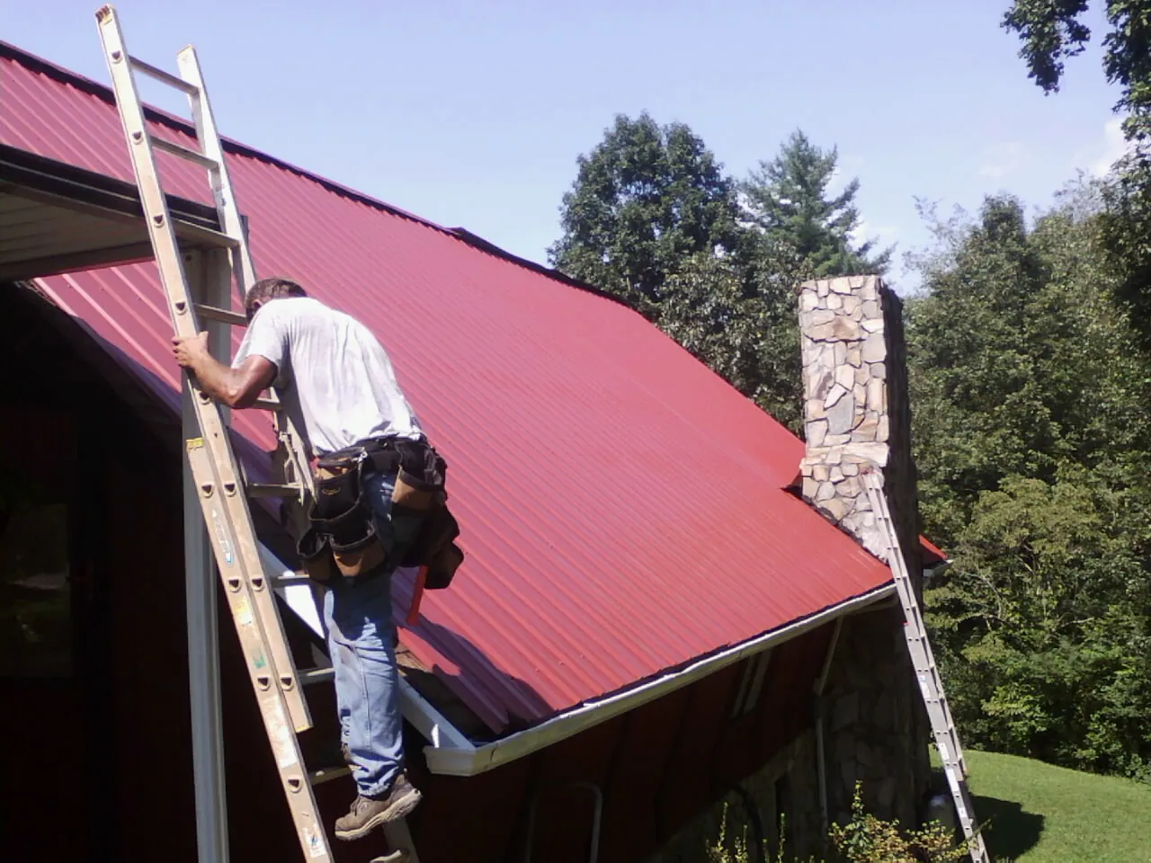 Roofer on a ladder beside a red metal roof and stone chimney — Soffit & Fascia Repair by Roofing Brothers in Ravenna